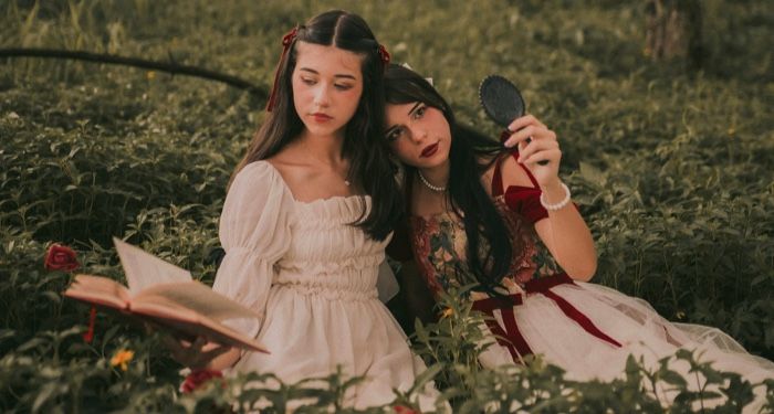 two fair skinned women dressed in a medievalfantastical theme sitting in a fieldone is reading a book.jpg.optimal