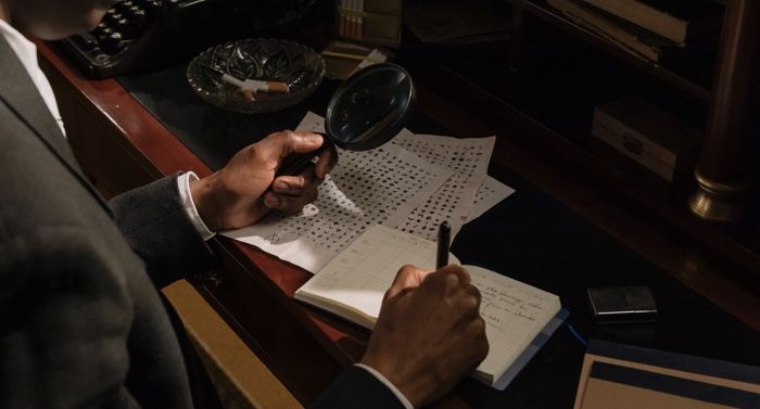 brown skinned hands holding magnifying glass and looking at papers on a desk.jpg.optimal